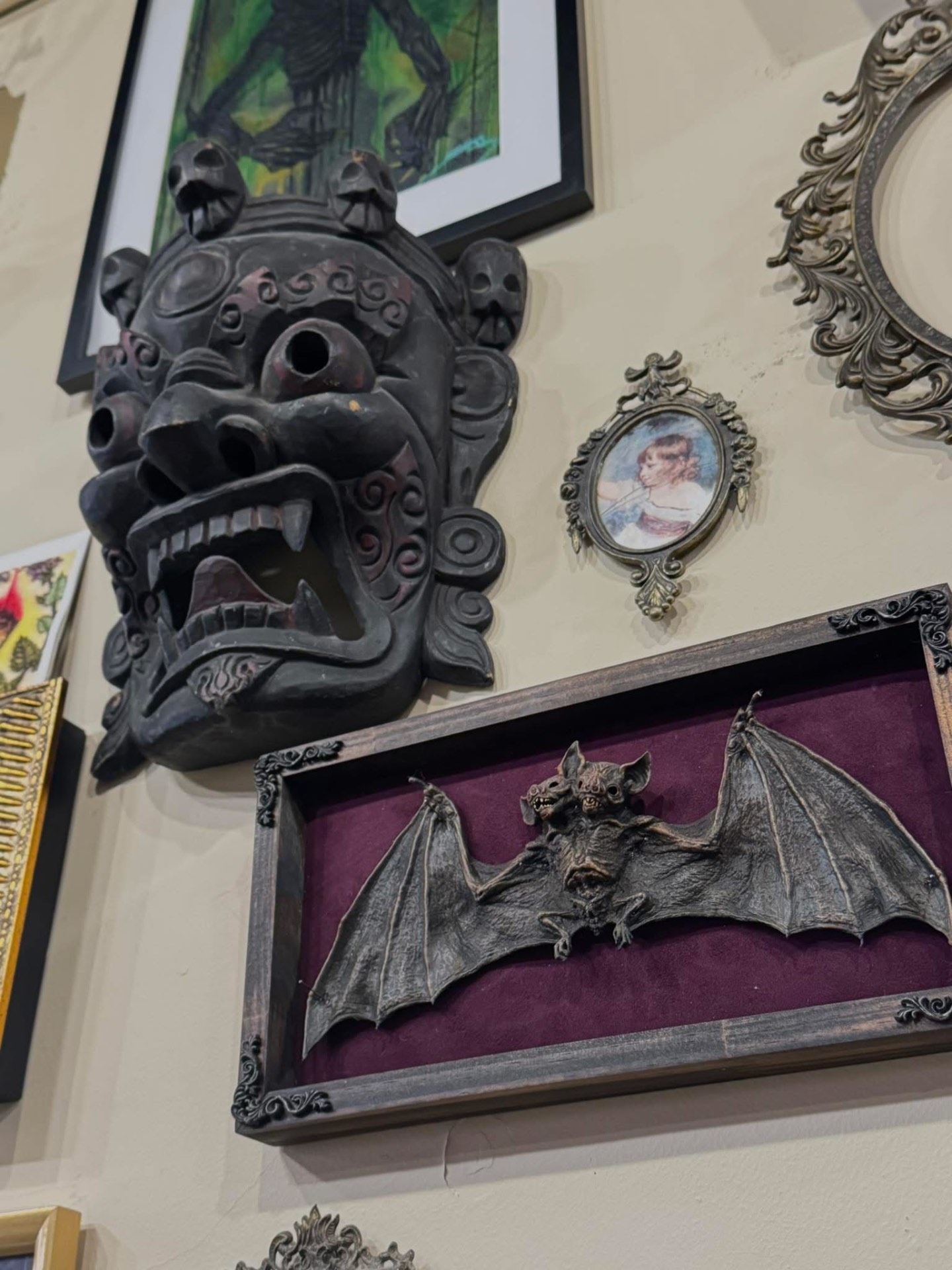 Framed bat specimen in a velvet shadow box beside a carved gothic demon mask on the studio wall